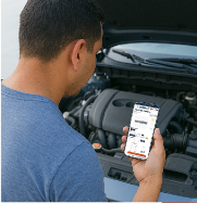 Person standing beside an open car hood, holding a smartphone displaying the BelMetric website
